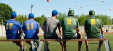 Fünf Jugendliche in Sportkleidung sitzen auf einer Bank eines Baseball-Spielfeldes und schauen einem Spiel zu